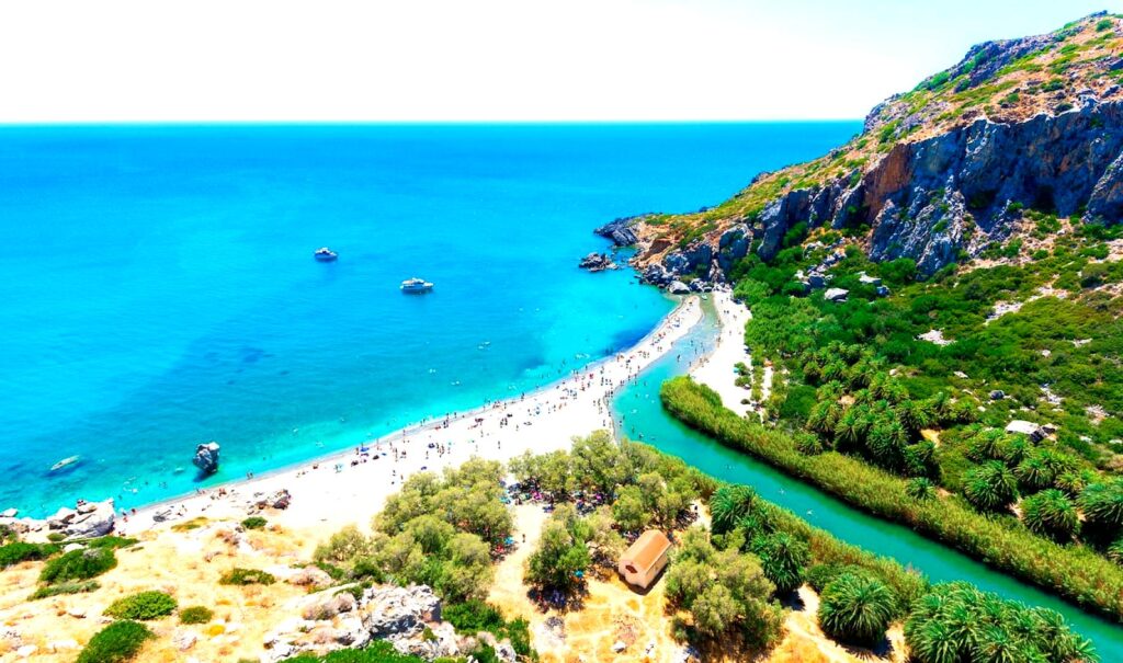 Preveli Beach is a scenic beach at the mouth of a river, lined with palm trees and surrounded by cliffs