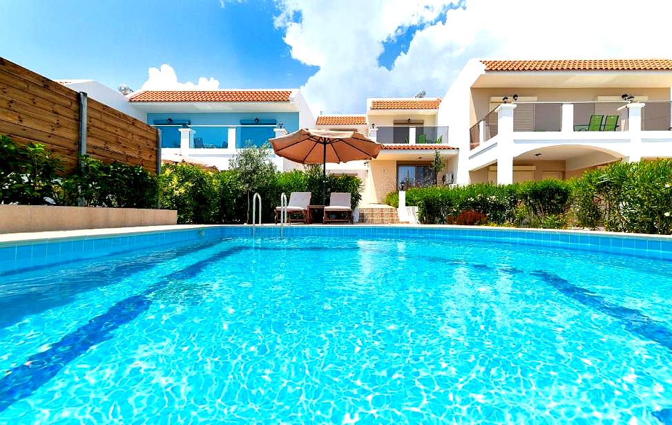 large outdoor pool with sunbeds and umbrella surrounded by greenery and modern villas